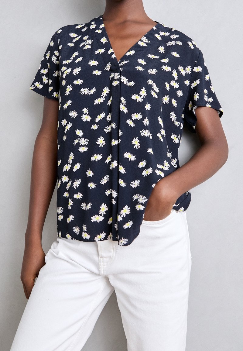 Personne portant un chemisier bleu marine à manches courtes avec des motifs de marguerites blanches et un pantalon blanc, une main dans la poche sur fond gris.
