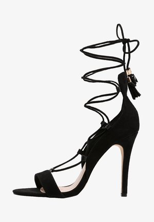 Des sandales à talons hauts en suede noir présentent un design à lanières, un bout ouvert, et un talon fin avec un laçage détaillé et des accents de franges.