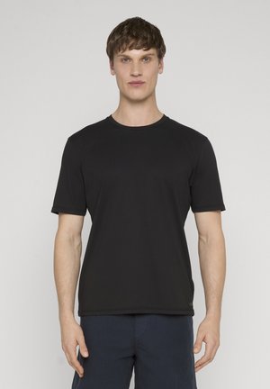 Joven con cabello corto y oscuro, de pie mirando al frente, vistiendo una camiseta negra de manga corta y pantalones oscuros, sobre un fondo claro.