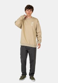 Beige sweatshirt with ribbed cuffs and hem, features a white logo on the chest. Paired with dark gray pants and green sneakers.