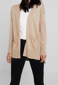 Beige lång kofta med ribbad struktur, öppen framdel och två framfickor, matchad med en vit topp och svarta byxor.