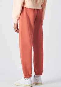 Pantalons de jogging confortables et amples en tissu doux, couleur terre cuite, avec des poignets élastiques. Le design comprend des poches latérales et un marquage discret.