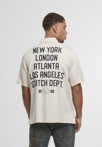 Camisa de manga corta de color crema claro con gran texto negro en la parte posterior que lista ciudades y "STITCH DEPT." Abajo hay un pequeño logo y el año "2002."