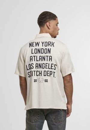 Light cream short-sleeve shirt with large black text on the back listing cities and "STITCH DEPT." Below is a small logo and year "2002."