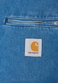 Modrá denimová látka s kapsou na zip. Obsahuje bílou nálepku se žlutým logem a textem "carhartt" v bordó pod ním.