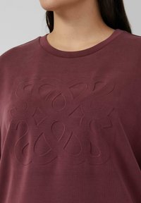 Bordowa koszulka z teksturowanym, wypukłym wzorem na przodzie. Okrągły dekolt, krótkie rękawy i miękka struktura tkaniny.