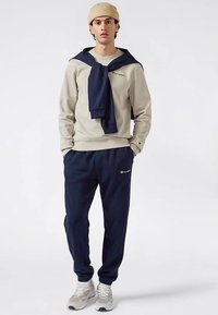 Beige sweatshirt with navy accents and logo, paired with navy pants. Model wears a navy shawl and beige beanie, and gray sneakers.