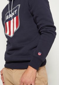 Homme portant un sweat à capuche GANT bleu marine avec un grand logo bouclier rouge et blanc sur la poitrine et un petit patch logo sur la manche, associé à un pantalon beige.
