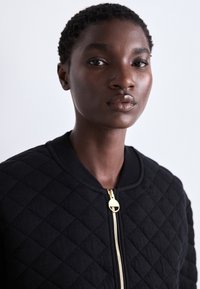 Casaco preto acolchoado com colarinho redondo e detalhes em zíper dourado. O tecido apresenta um padrão de diamante texturizado e uma aparência suave e macia.