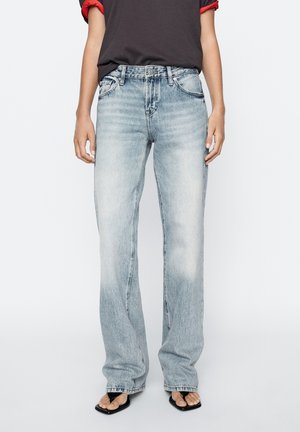 Person iført lyseblå straight-leg jeans, sorte sandaler med firkantet tå og en sort t-shirt med røde optrukne ærmer.