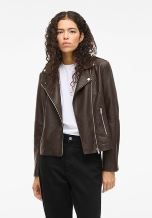 Bruine leren motorjack met ritssluiting aan de voorkant, zilverkleurige hardware en een schuin kraag. G gedragen over een witte shirt en gecombineerd met zwarte broek.