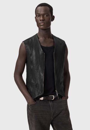 Ung mand iført en sort lædervest uden ærmer over en sort skjorte og mørke jeans med hænderne i lommerne foran en ensfarvet baggrund.