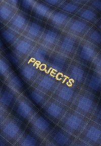 Kék kockás anyag sárga hímzett szöveggel: "PROJECTS." A textúra puha, szövött felszínű, finom rácsmintával.