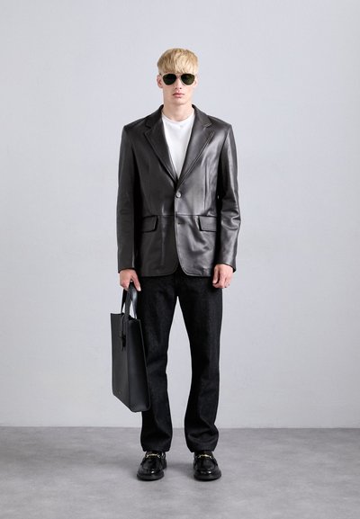Blond man wearing black leather blazer, white shirt, black pants, black loafers with gold detail, sunglasses, holding black leather tote bag.