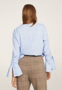 Chemise bleu clair à rayures avec des détails de cravate aux manches, associée à un pantalon à carreaux dans des tons atténués. Texture en tissu lisse, coupe décontractée.