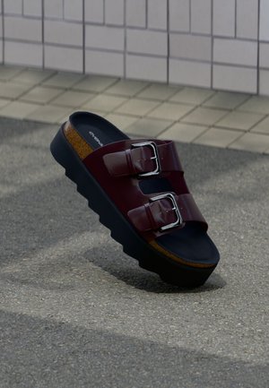 Sandale roșu închis cu două catarame argintii și talpă groasă neagră, așezată pe paviment gri lângă un perete placat cu gresie.