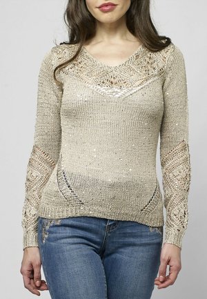 Kobieta ubrana w beżowy, dziergany sweter z koronkowymi wzorami na klatce piersiowej i rękawach, w połączeniu z niebieskimi dżinsami.