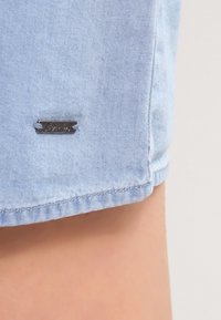 Vaaleansininen farkkukangas sisältää metallilapun, jossa on kaiverrettu "Denim". Se on sijoitettu lähelle helmaa, ja reunat ovat hieman risat.
