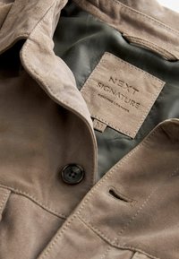 Pruunist sametist jakki nupuväravaga, millel on õmmeldud sildiga etikett, millel on kirjas "NEXT SIGNATURE GENUINE LEATHER" kontrastsetes tekstuurides.