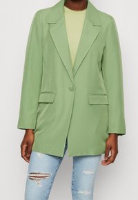 Blazer verde con colletto a lancia, chiusura con un solo bottone e tasche frontali. Abbinato a una maglietta chiara e jeans blu strappati.