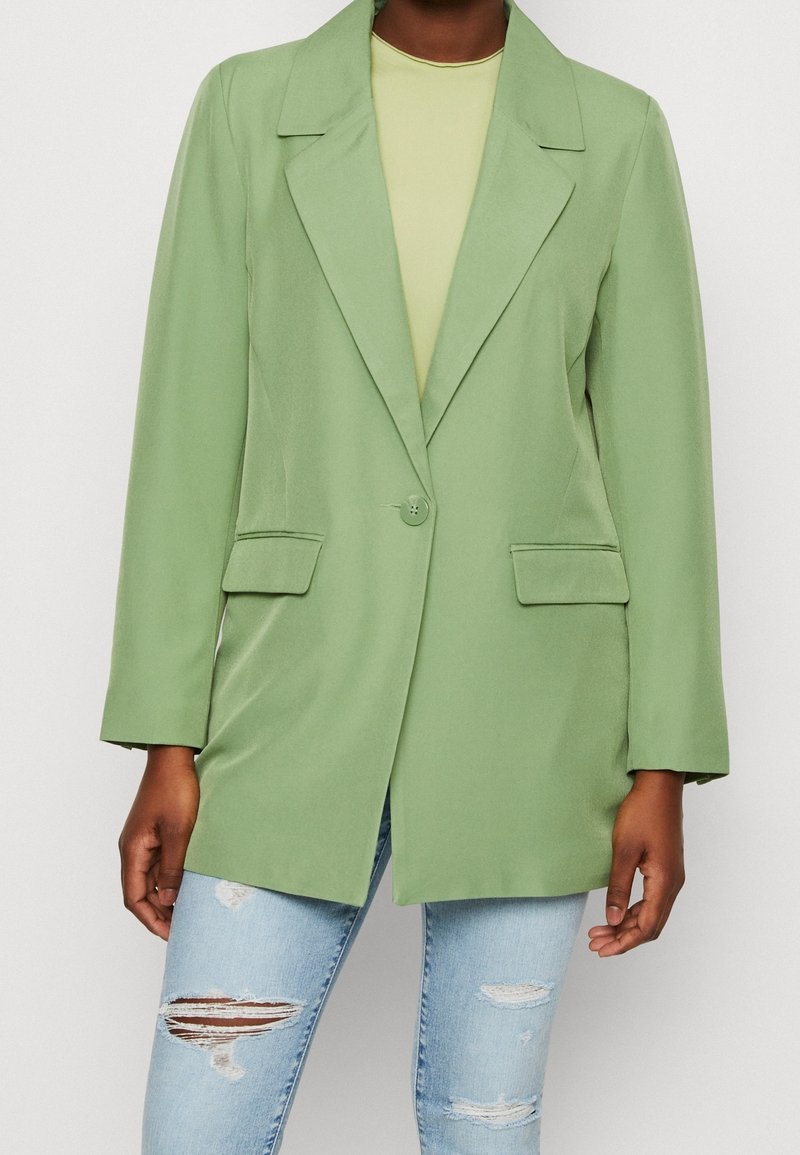 Blazer verde con colletto a lancia, chiusura con un solo bottone e tasche frontali. Abbinato a una maglietta chiara e jeans blu strappati.