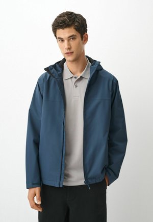 Junger Mann trägt eine blaue Kapuzenjacke über einem hellgrauen Poloshirt und steht vor einem schlichten weißen Hintergrund.