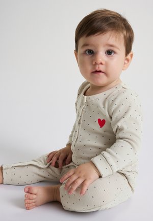 Tout-petit aux cheveux bruns assis, portant un pyjama crème à pois avec un cœur rouge sur la poitrine, regardant légèrement sur le côté sur un fond blanc.