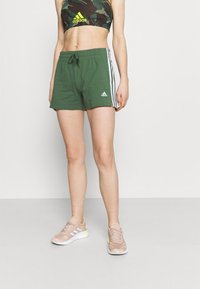 Bermudas deportivas de algodón verde con franjas laterales blancas y una cinturilla elástica, combinadas con zapatillas beige con detalles en blanco.