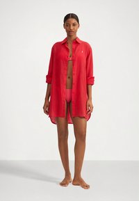 Red linen shirt dress with a button-down front, rolled sleeves, and a loose fit, worn over a matching pink bikini. Smooth texture.