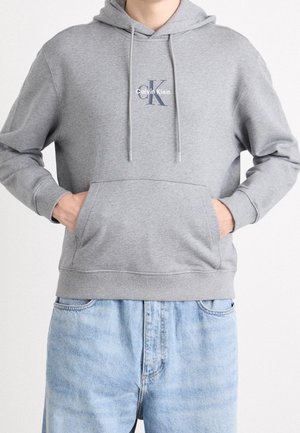 Osoba ubrana w szarą bluzę z kapturem Calvin Klein z przednią kieszenią i troczkami, w połączeniu z jasnoniebieskimi dżinsami, z rękami w kieszeniach.