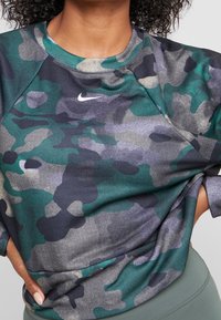 Szara i zielona bluzka z okrągłym dekoltem w kamuflażowy wzór, wykonana z gładkiego materiału, z małym białym logo Nike na górze po lewej stronie.