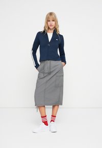 Námornícky zip-up kabát s bielymi postrannými pruhmi, šedá sukňa po polovicu lýtok s vreckami, červené a biele pruhované ponožky a biele tenisky.