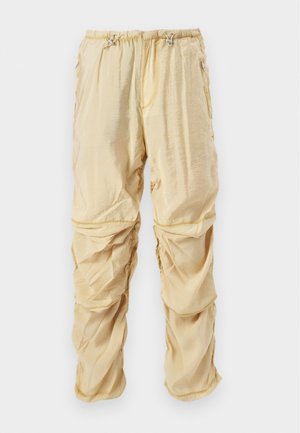 Calças cargo de nylon de um tom bege claro, com cordões ajustáveis na cintura e bolsos com fecho, projetadas com detalhes de tecido franzido e em camadas.