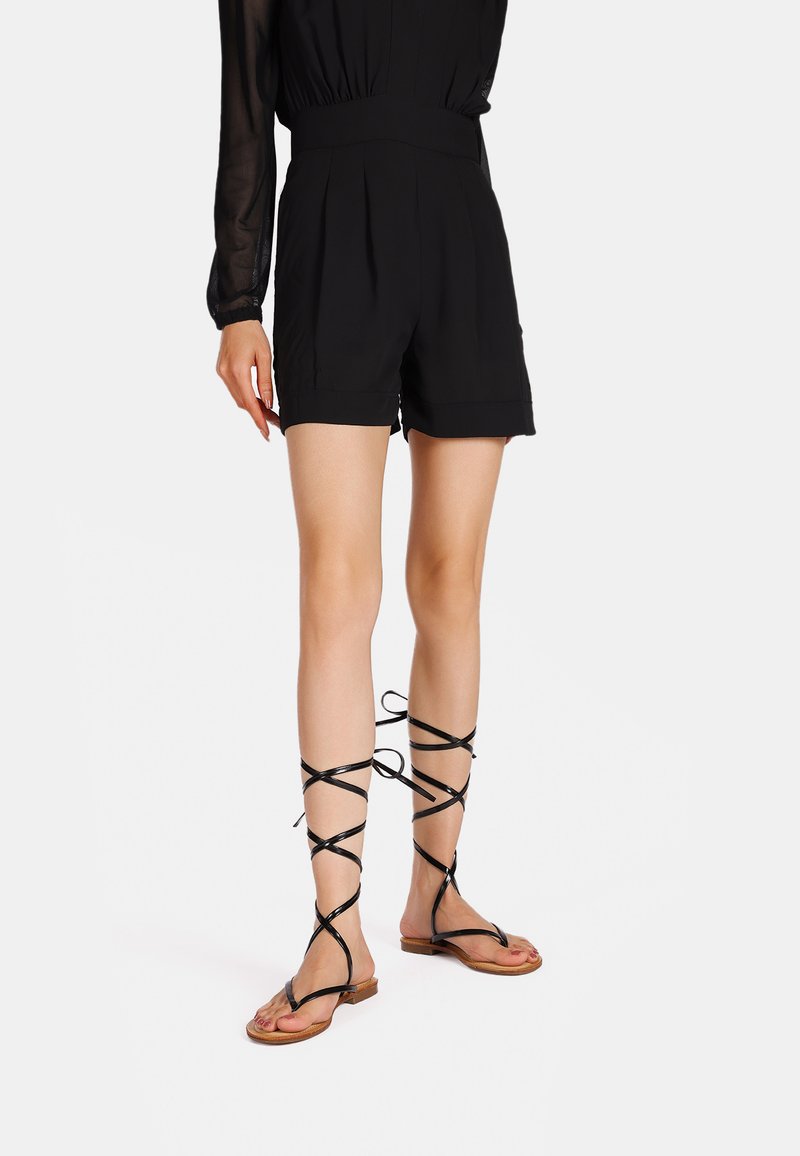 Shorts neri con pieghe e vita alta, abbinati a sandali neri con cinturini alla caviglia, dotati di suole piatte e cinturini sottili.