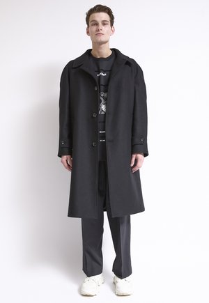 Junger Mann, der vor einem weißen Hintergrund steht und einen langen schwarzen Mantel, schwarze Hosen, weiße Turnschuhe und ein schwarzes Sweatshirt mit Grafik trägt.