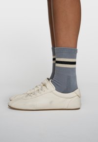 Les baskets en cuir blanc présentent une finition texturée avec un bout lisse et un devant à lacets. Des chaussettes grises avec des rayures blanc crème et noires sont portées.