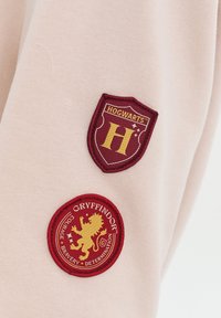 Ružová textília oblečenia s dvomi vyšívanými nášivkami: štít s nápisom "HOGWARTS" a leva s nápisom "GRYFFINDOR."