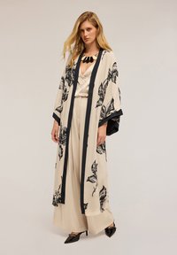 Dlouhý béžový kimono s černým květinovým potiskem, černým lemem a širokými rukávy, nošený přes světle zabarvený, splývavý top a široké kalhoty.