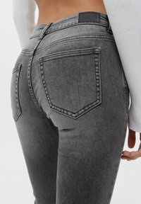 Jean en denim gris avec une coupe ajustée, des poches arrière et une finition texturée. La ceinture présente un accent noir.