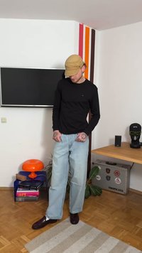 Black long-sleeve shirt, light blue wide-leg jeans, brown shoes, light tan cap; standing in a room with various colored accents and decor.