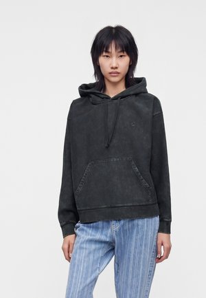 Giovane donna con capelli neri che indossa una felpa nera sbiadita e jeans azzurri a righe, in piedi davanti a uno sfondo bianco semplice.
