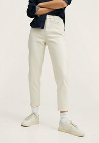 Jeans color crema con un ajuste slim, cintura alta y tobillos recortados. Combinados con zapatillas ligeras y calcetines blancos hasta el tobillo.