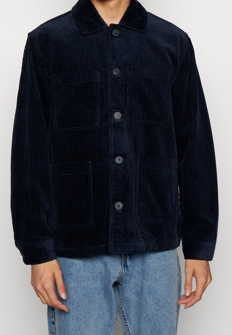 Chaqueta de pana azul marino con cuello, cierre de botones y dos bolsillos frontales. Tejido texturizado con estrías verticales y corte relajado.