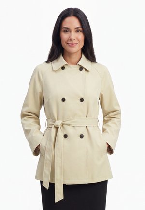 Frau mit langen dunklen Haaren, die einen beigen, doppelt geknöpften Trenchcoat trägt, in der Taille gebunden, Hände in den Taschen, lächelnd vor weißem Hintergrund.