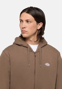 Bruine zip-up hoodie met een trekkoordkap, grote zijzakken en een klein wit logo op de borst. Zacht materiaal met een gladde textuur.