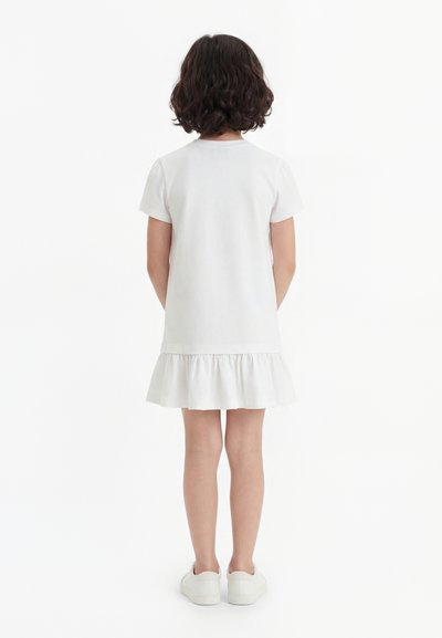 Enfant aux cheveux courts et bouclés, debout de dos, portant une robe blanche à manches courtes avec un ourlet à volants et des baskets blanches.