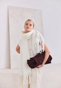 Witte wollen sjaal met franjerand, gecombineerd met een donkerbruine suède tas. Op de achtergrond een lichtgekleurde muur met textuur.
