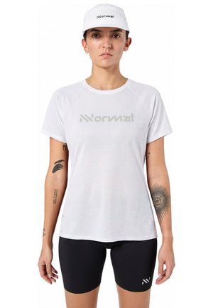 Person trägt weiße Kappe und weißes kurzärmeliges Hemd mit "Normal"-Logo, schwarze Shorts mit kleinem Logo, steht vor weißem Hintergrund.