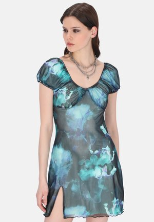 Femme portant une robe transparente à manches courtes avec un motif floral dans des tons de bleu et de vert, avec une fente à l'avant et des colliers argentés superposés.