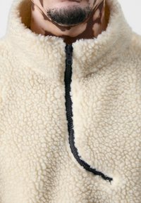 Cremfarbener Fleece-Jacke mit halbem Reißverschluss, der eine weiche, strukturierte Oberfläche und einen schwarzen Reißverschlussakzent aufweist.